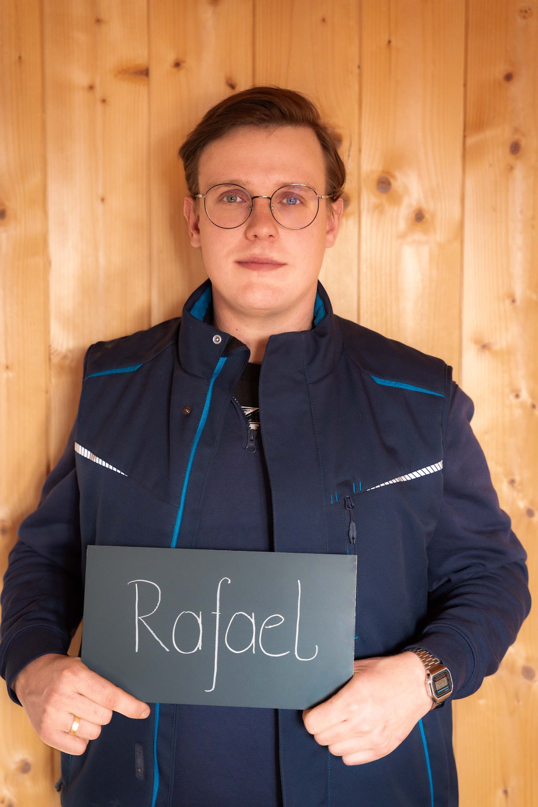 Ein Mann hält vor einer Holzwand ein Schild mit der Aufschrift „Rafael“.