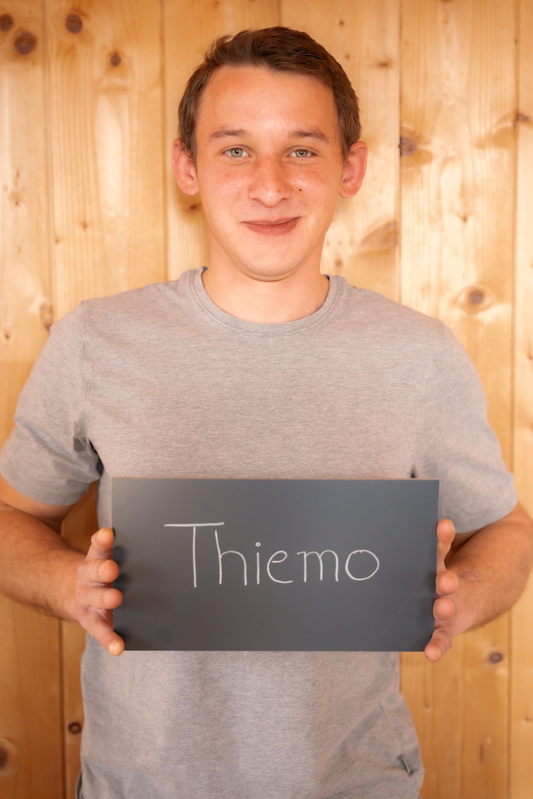 Ein junger Mann hält vor einer Holzwand ein Schild mit der Aufschrift „Thiemo“.