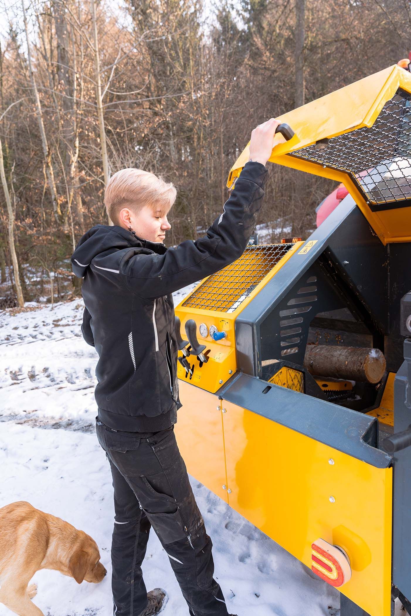 Ein Mädchen öffnet im Schnee die Motorhaube eines gelben Fahrzeugs.