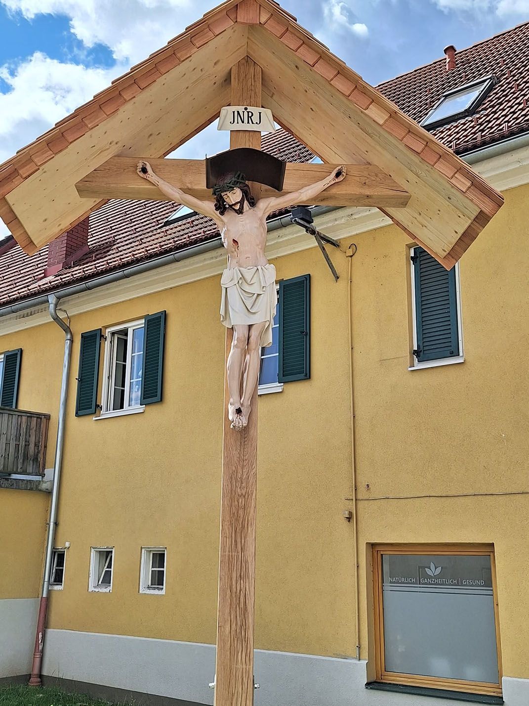 Vor einem gelben Gebäude steht ein Holzkreuz mit Jesus darauf.