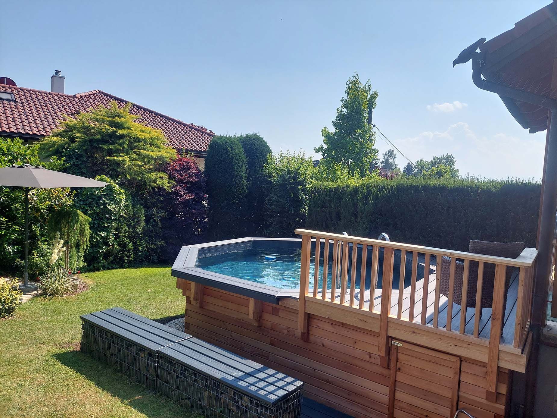 Hinterhofpool mit Holzdeck und Geländer, grünem Rasen, Bank und umgebendem Grün.