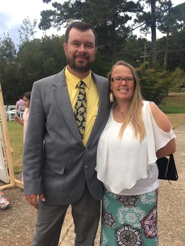 Two people standing outdoors, one in a gray suit and yellow tie, the other in a white top and patterned skirt.