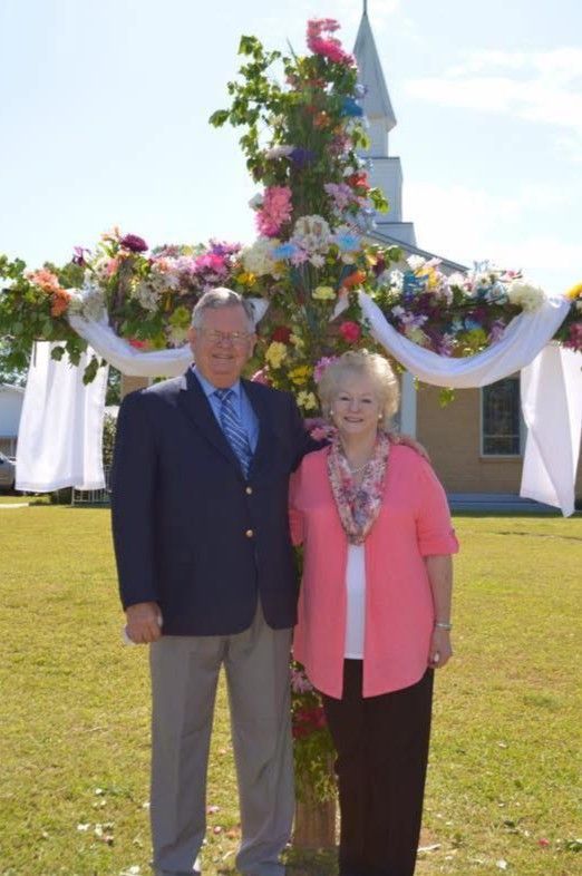 Two people standing on grass in front of a flower-covered arch and church, dressed for a formal outdoor event