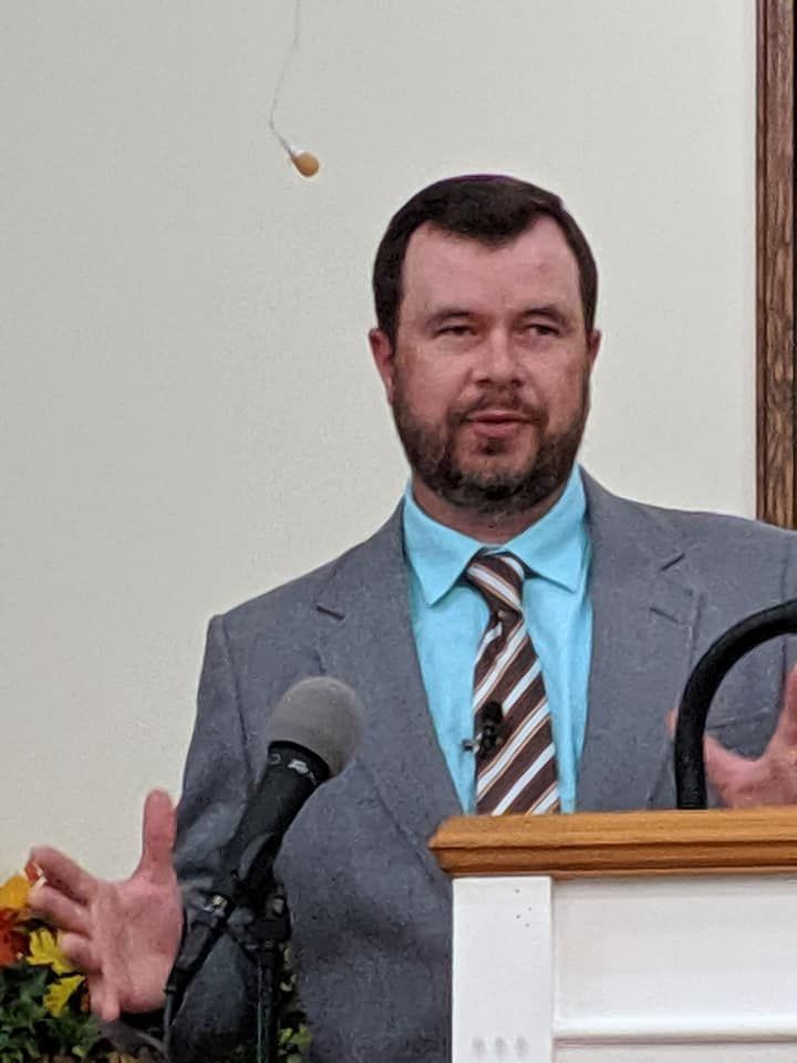 Man speaking at a podium in a gray suit and blue shirt, with a microphone in front of him
