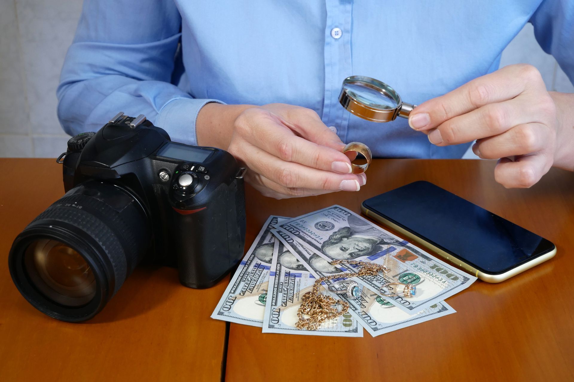 Pawnbroker inspects a gold ring with a magnifier beside cash, jewelry, a camera, and a smartphone