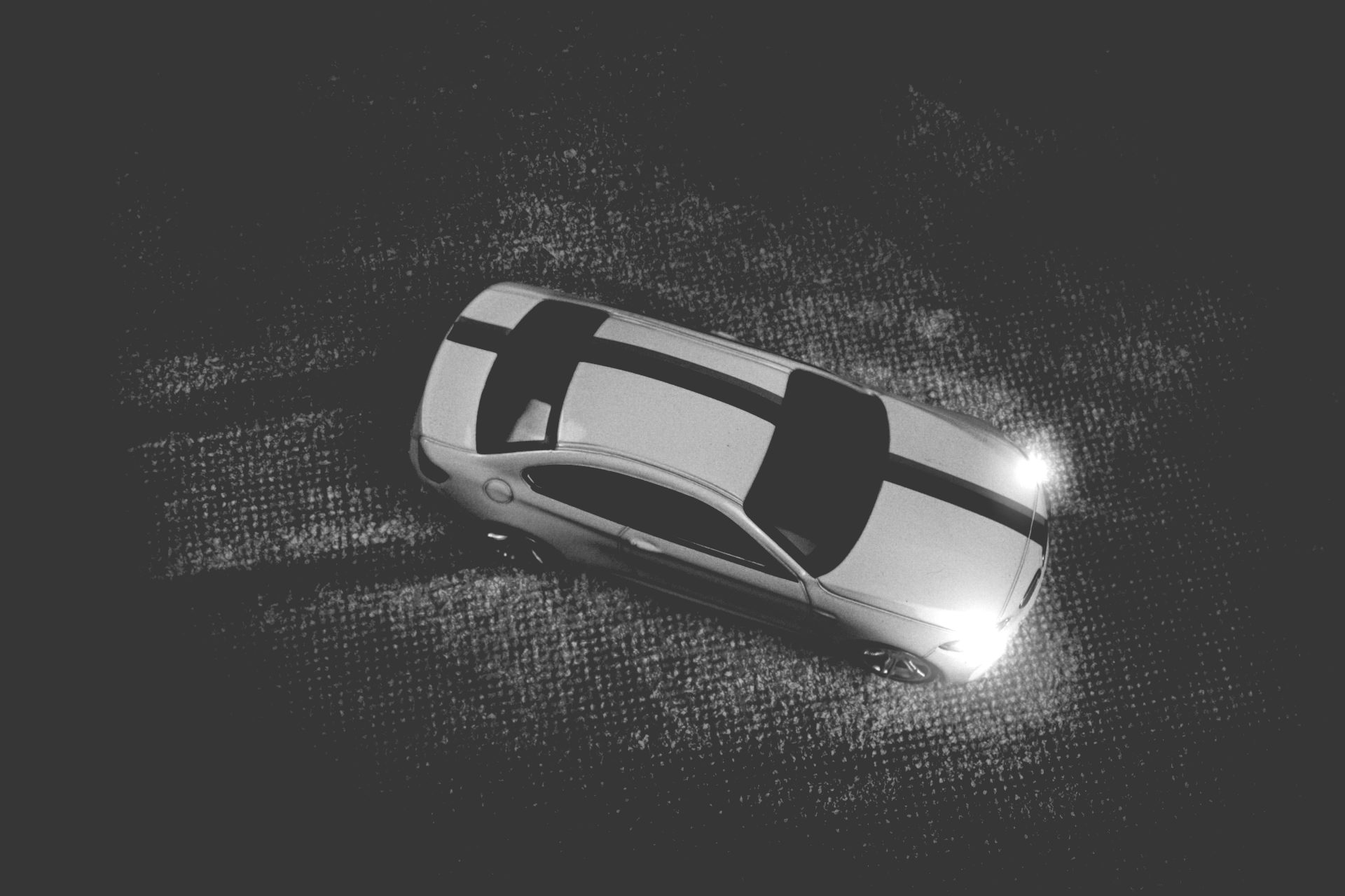 Top-down view of a white car with black stripes driving on a dark surface, headlights on.