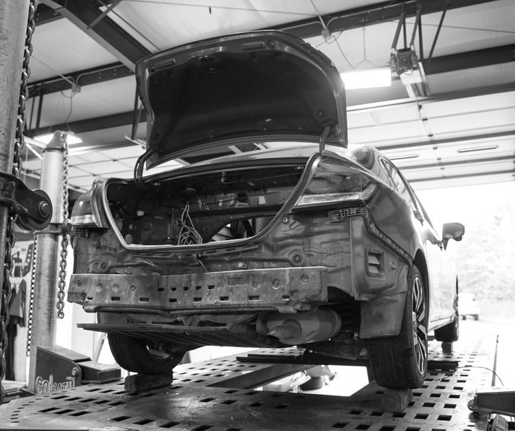 Rear of a damaged black car on a lift in a repair shop, trunk open, bumper removed.