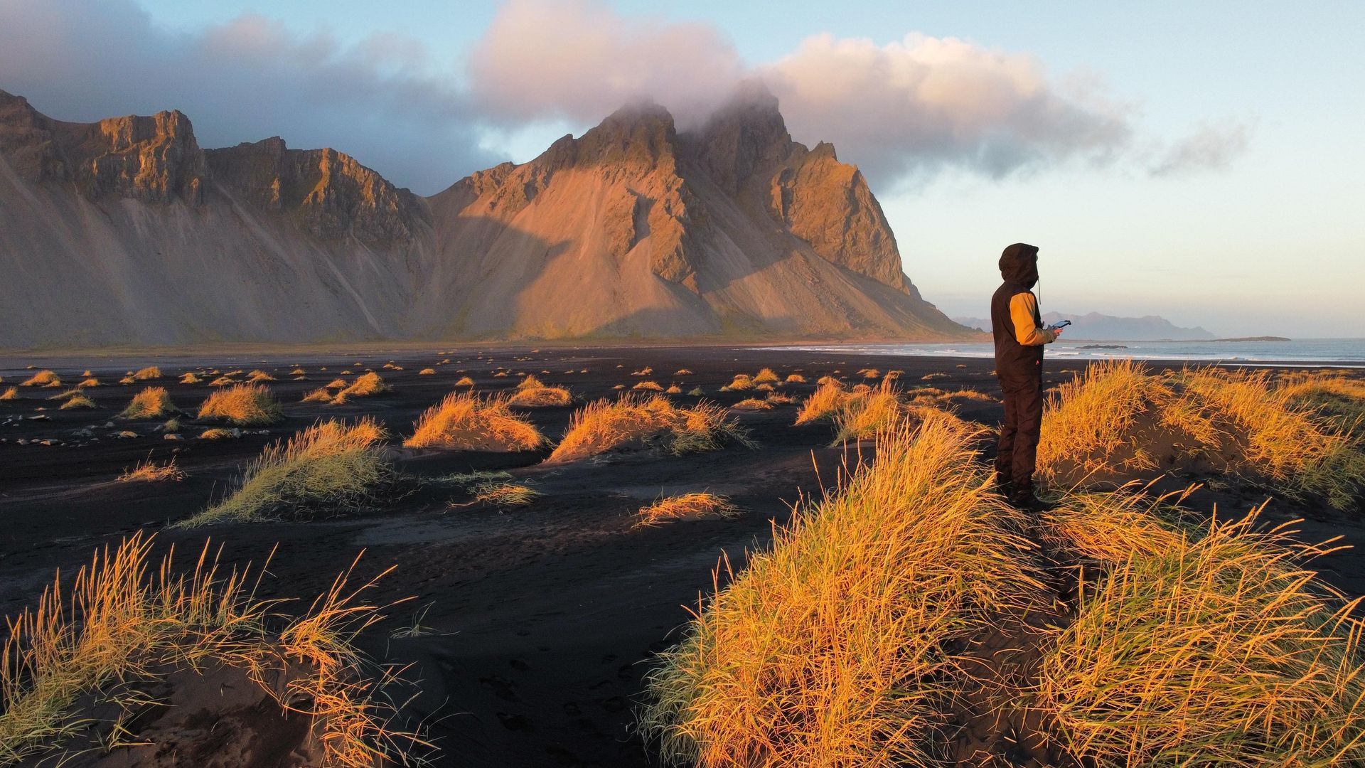 Eine Person steht an einem schwarzen Sandstrand, im Hintergrund Berge, im Vordergrund goldenes Gras.