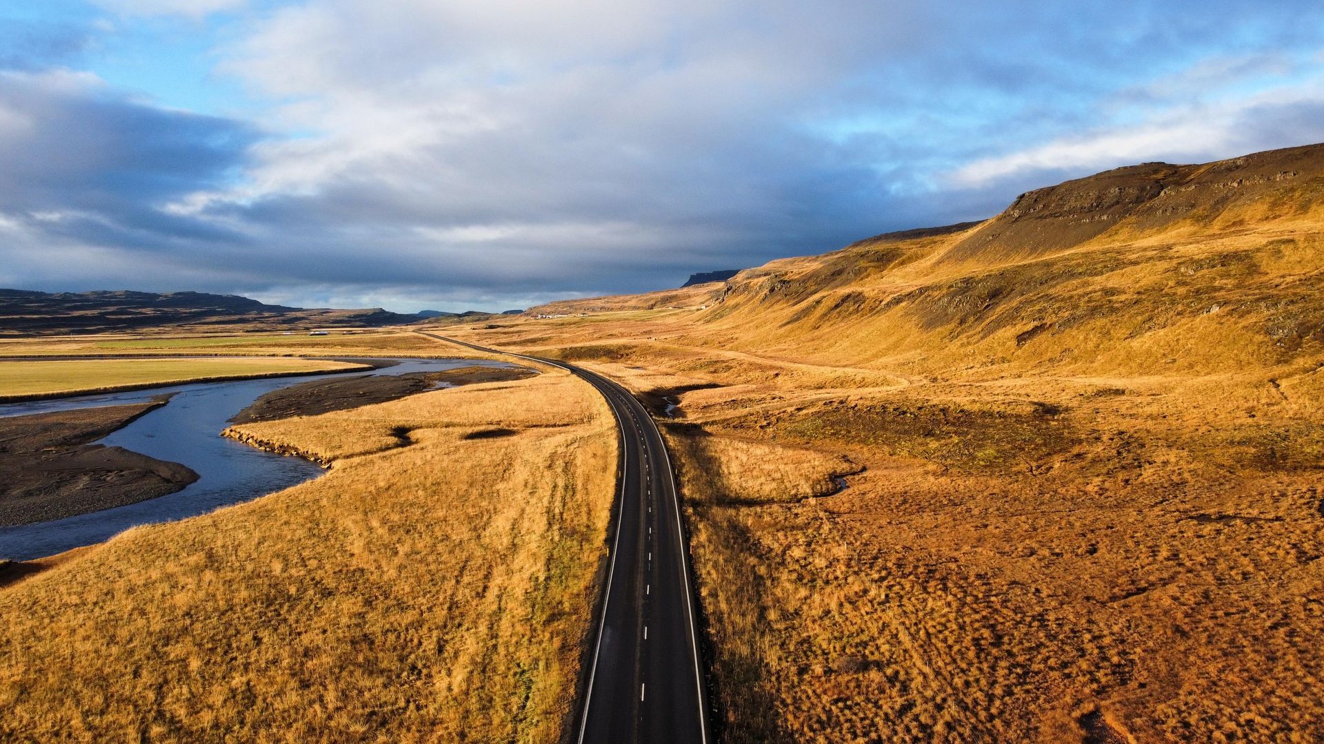 Die Strasse schlängelt sich durch eine goldene Landschaft, neben einem Fluss unter einem bewölkten Himmel.