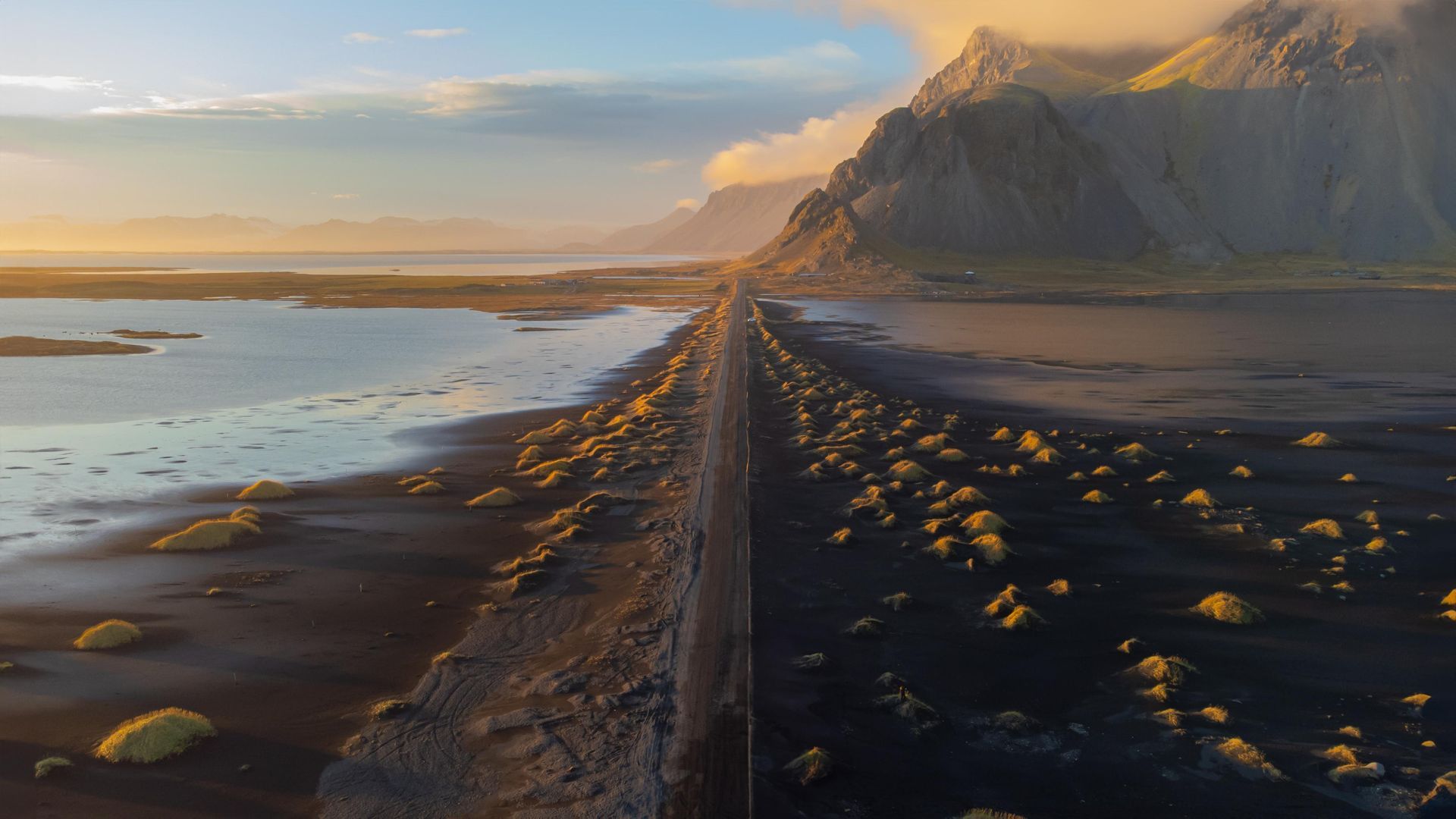 Eine Küstenstrasse erstreckt sich zwischen Wasser und schwarzem Sand, im Hintergrund erheben sich in goldenes Licht getauchte Berge.