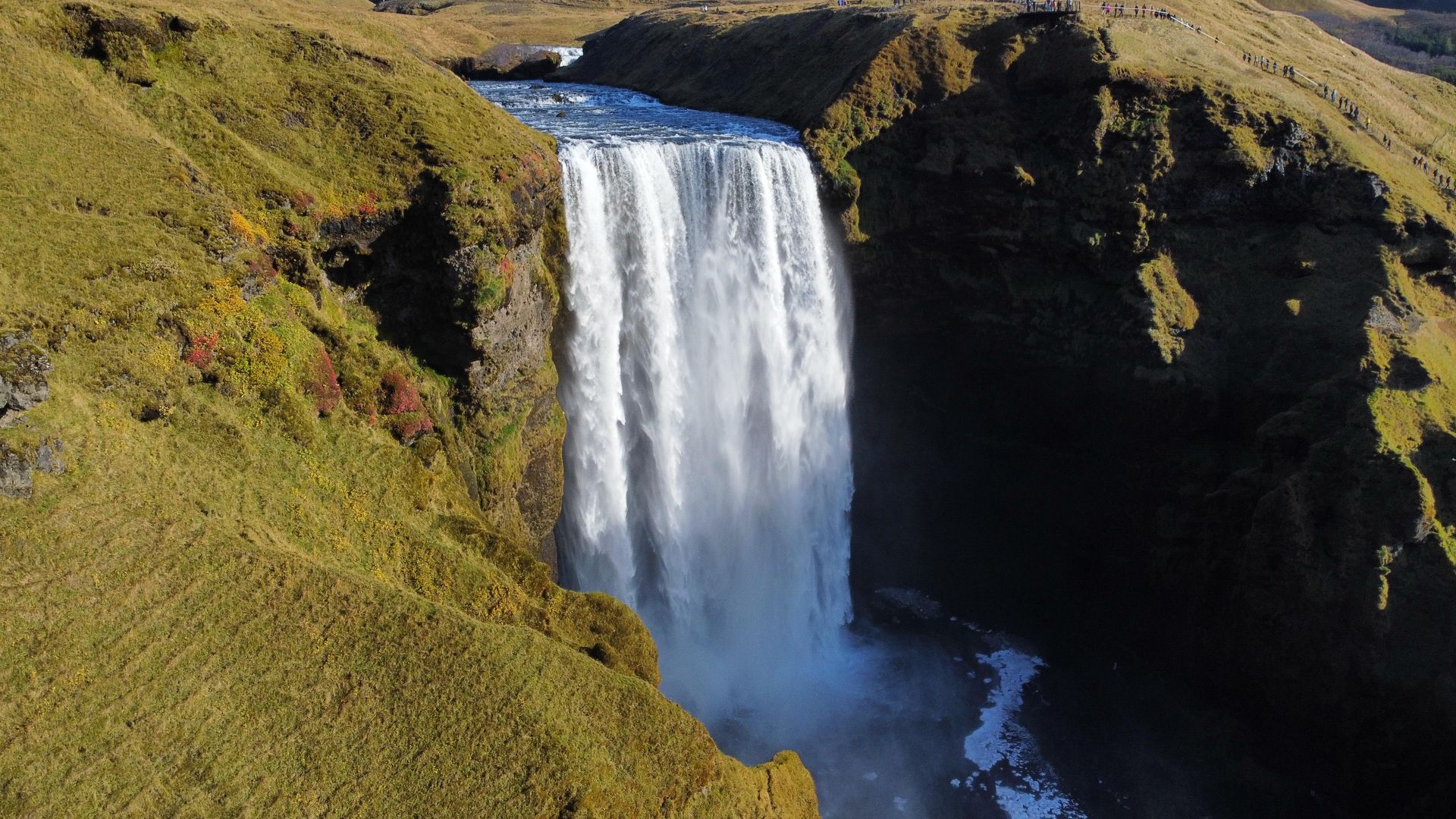 Ein Wasserfall stürzt über eine Klippe, umgeben von grasbewachsenen Hügeln unter sonnigem Himmel.