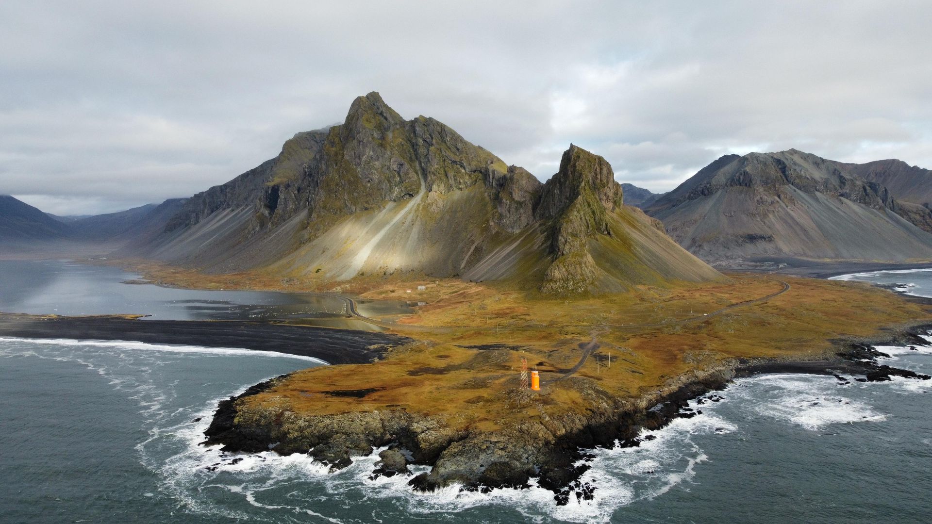 Karge Insel mit einem gelben Leuchtturm im Meer, überragt von zerklüfteten Bergen unter einem bewölkten Himmel.