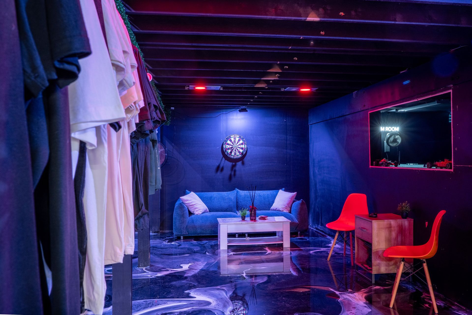 Room with clothing racks, blue couch, dartboard, and red chairs under dark lighting.