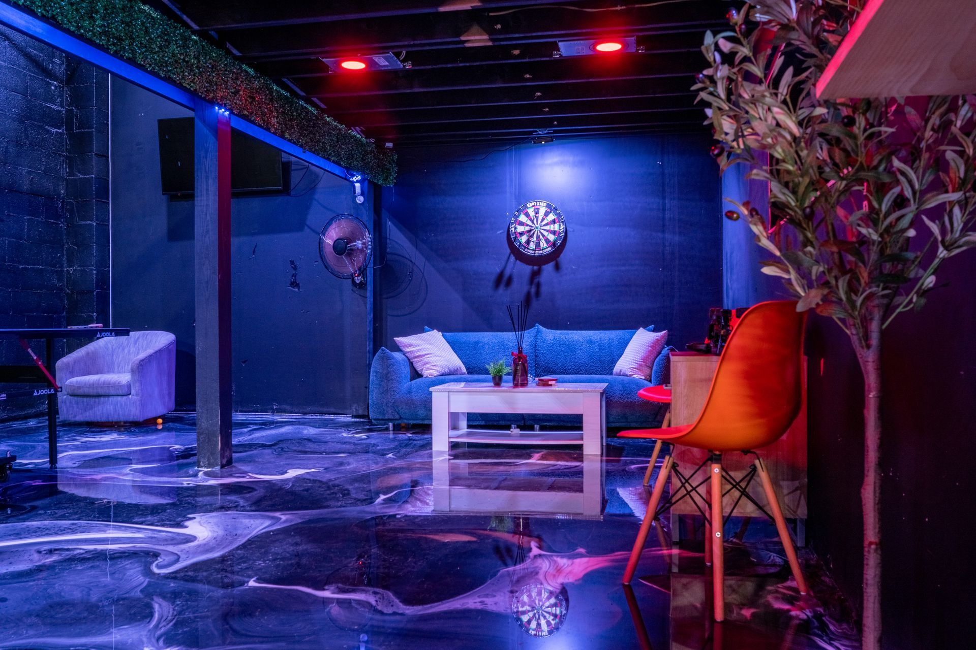 Dark room with blue and red lighting, couches, dartboard, and abstract floor pattern.