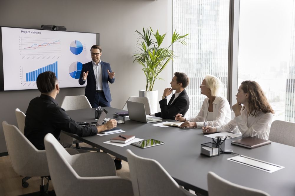 Man presenting data charts on screen to seated colleagues at a conference table.