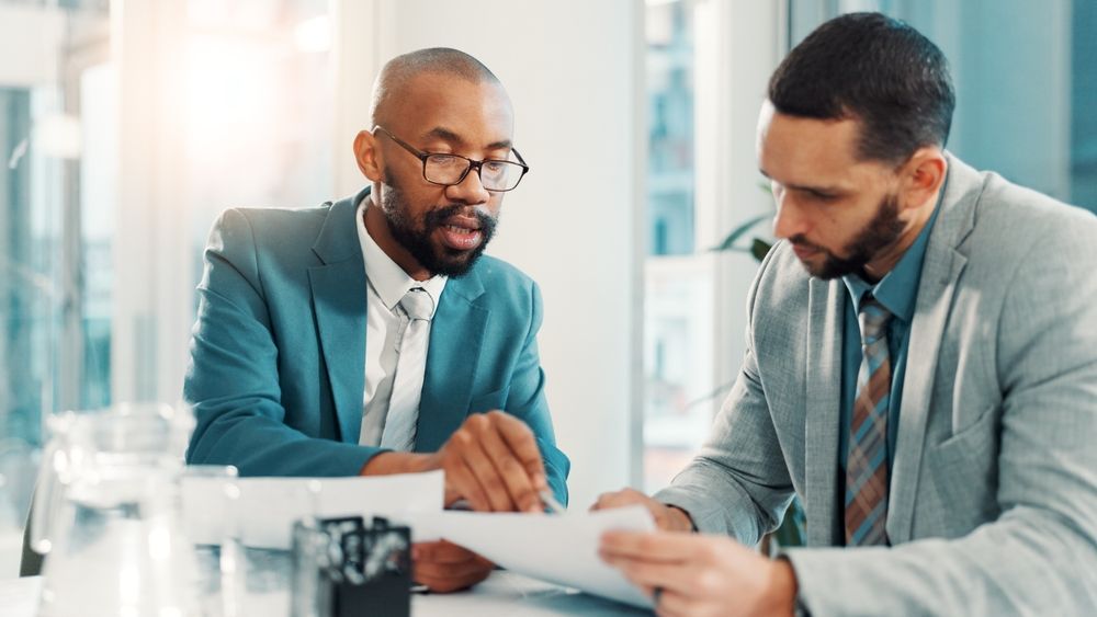 Two men in suits reviewing documents at a table; one points to the papers.