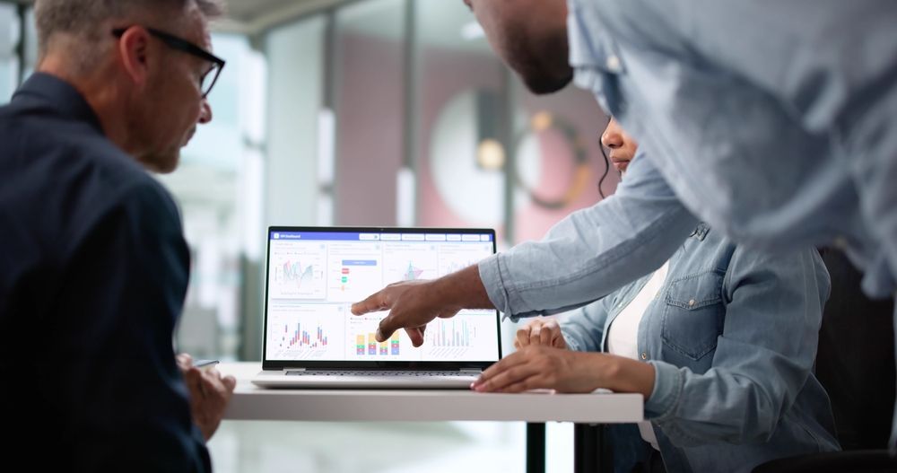 Three people reviewing data on a laptop screen, pointing and discussing, in an office setting.