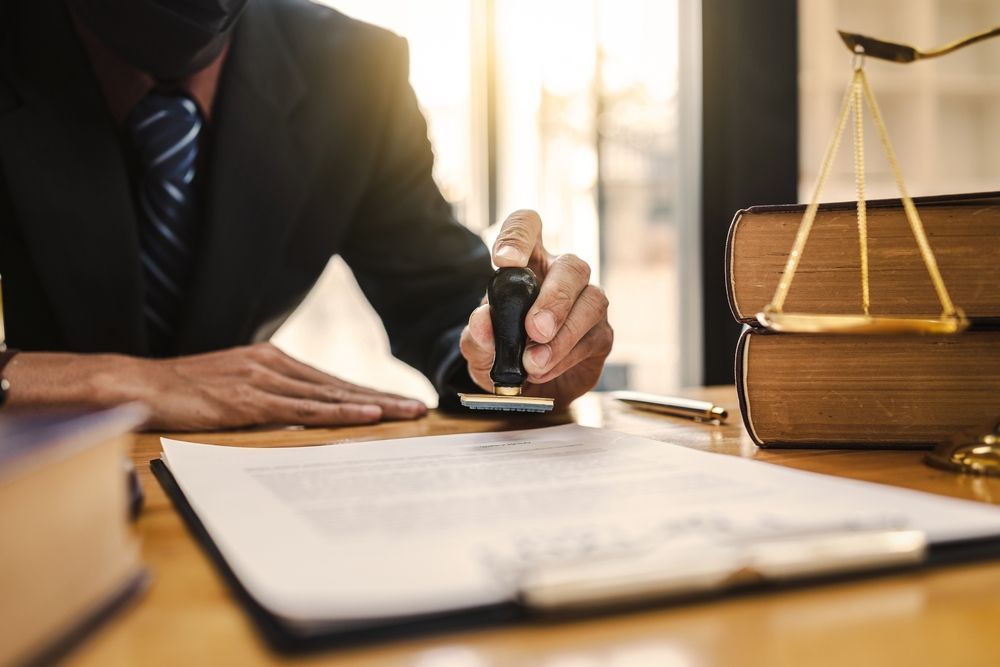 A person in a suit stamps a document on a desk, next to law books and a balance scale.