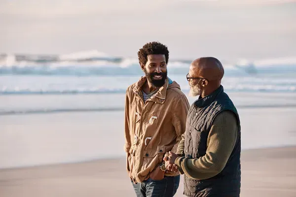 Two people walking and talking on a beach, with ocean waves in the background.