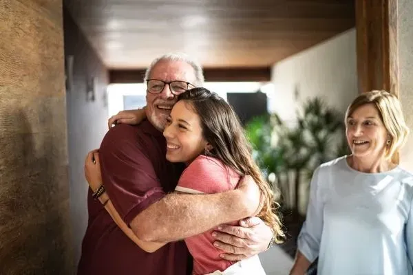 Two people warmly hugging in a hallway, with a smiling woman standing nearby.