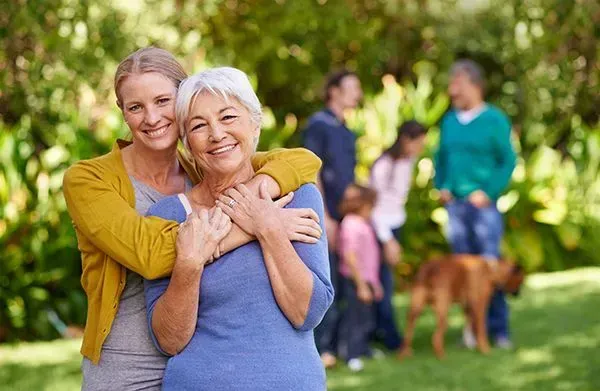 Two people smiling and hugging in a sunny park, with others and a dog blurred in the background