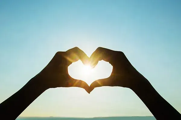 Hands forming a heart shape around the sun at sunset against a blue sky