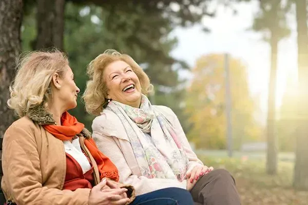 Two friends laughing on a park bench in autumn, with soft sunlight and trees in the background