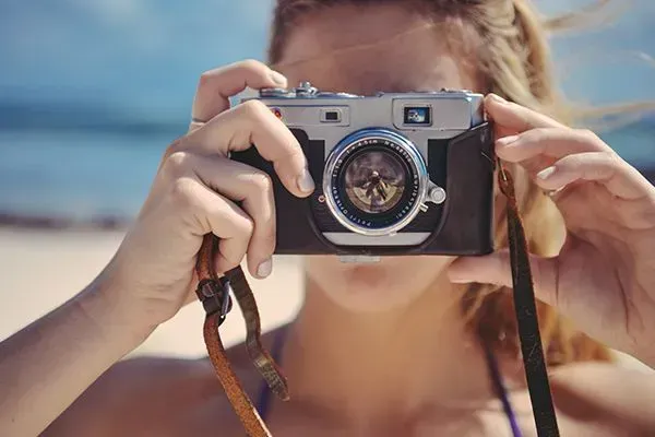 Person holding a vintage camera to their face on a sunny beach, with ocean blurred in the background