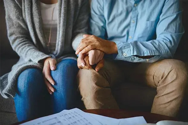 Two people seated close together, holding hands on a couch with papers in the foreground.
