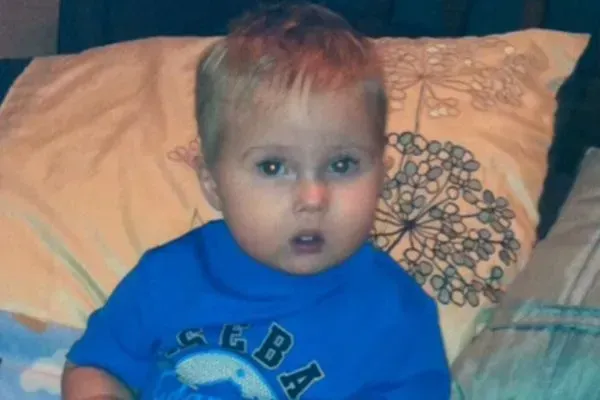 Toddler in a blue shirt sitting on a bed with patterned pillows, looking at the camera.