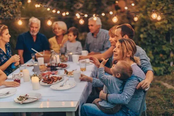 Family gathered around an outdoor dinner table under string lights, sharing food and conversation at dusk.