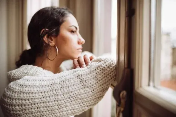 Woman in a cream sweater gazes out a window, resting her chin on her hand.