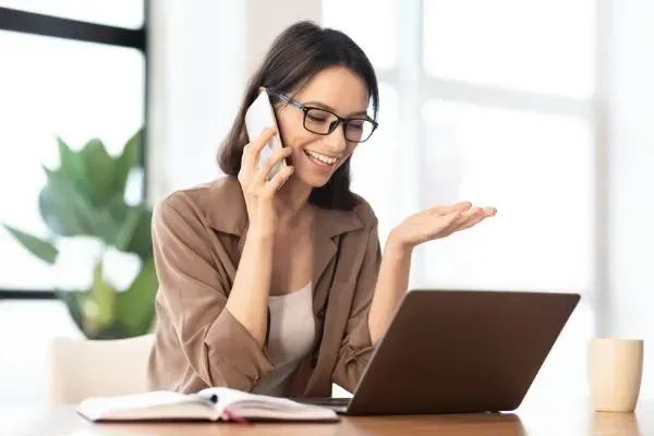 Woman on phone beside laptop, smiling at a desk in a bright home office.