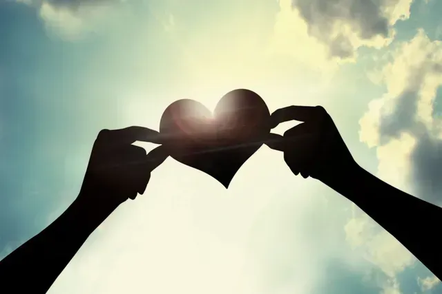 Hands holding a black heart silhouette against a bright sky with sun flare