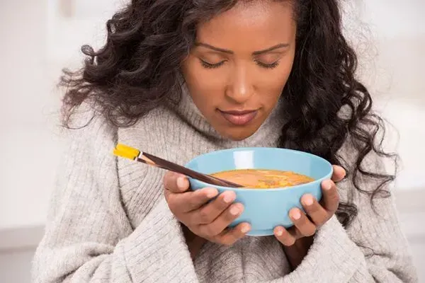 Woman in a gray sweater holding a blue bowl of soup with a spoon, looking down.