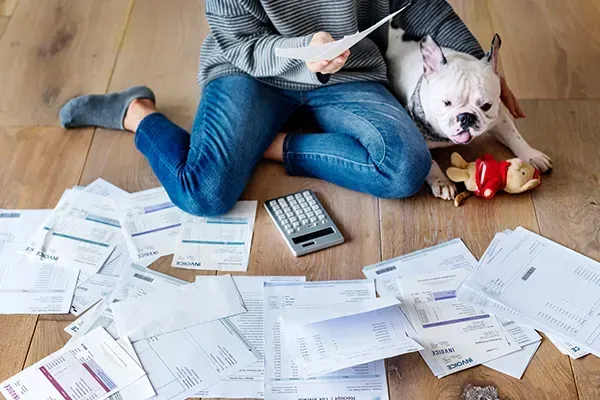 Person sitting on a wooden floor with scattered bills and a calculator beside a bulldog