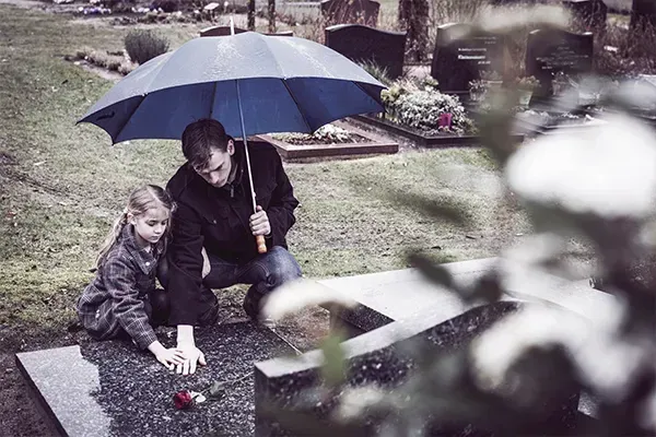 Two people sit under a blue umbrella by a graveside, placing flowers on a grave in the rain.