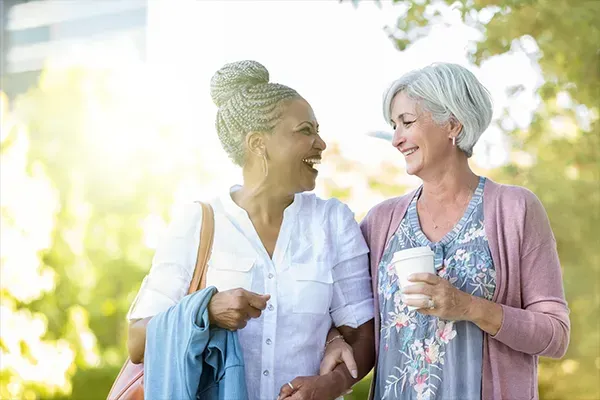 Two women smiling and walking outdoors, one holding a drink and the other a jacket.