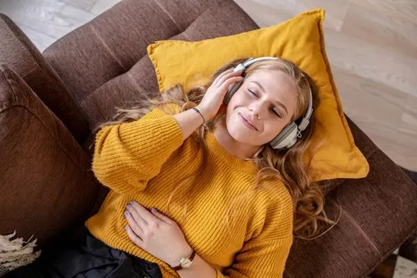 Woman in a mustard sweater relaxing on a brown couch with a yellow pillow, wearing headphones and smiling
