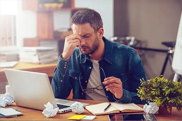 Stressed man at desk rubbing his eyes beside a laptop, papers, and coffee in a bright office