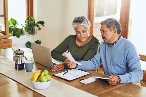 Two people reviewing documents at a kitchen table beside a laptop and fruit bowl