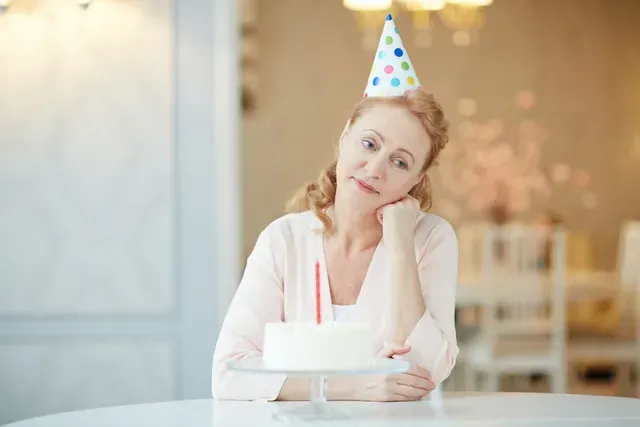 Woman wearing a party hat holding a cake, smiling thoughtfully at a decorated table.