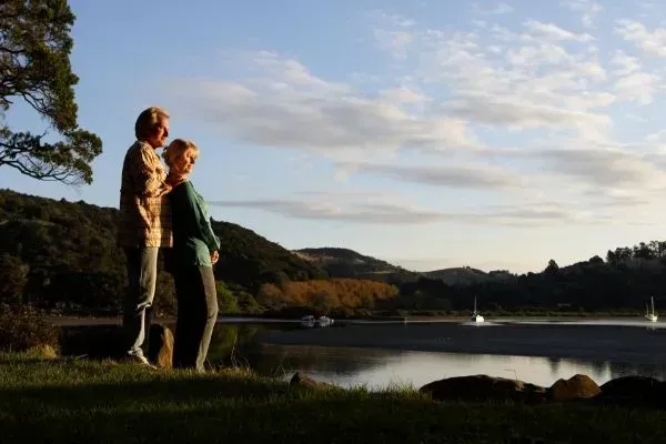 Two people standing by a lakeside at sunset, looking across the water to distant hills.