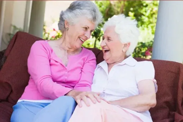Two smiling women sit together on a couch, chatting outdoors in bright sunlight.