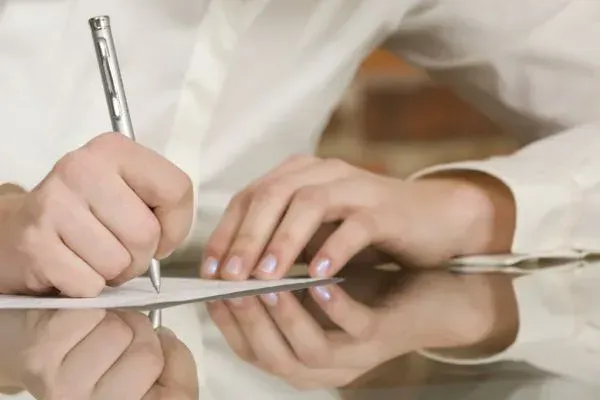 Hands writing on paper with a pen at a reflective table surface