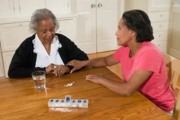 Two women sit at a table, one comforting the other, with a pill organizer and water glass nearby.