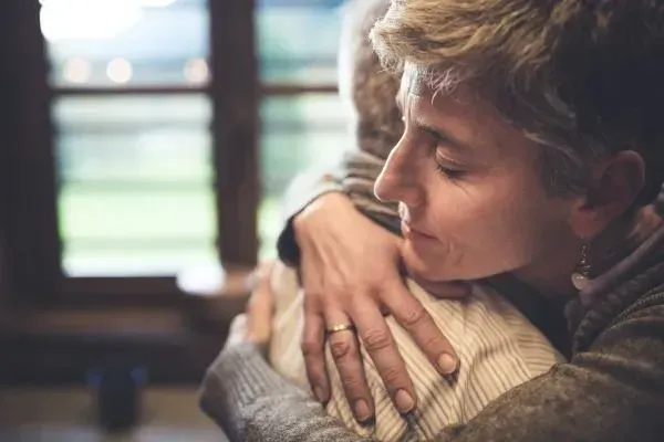 Two people hugging indoors, one with eyes closed, in a warm, softly lit room
