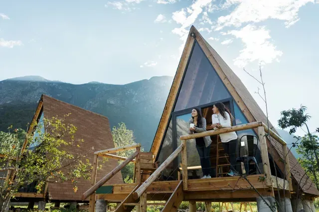 Two people on a wooden A-frame cabin balcony with mountain backdrop
