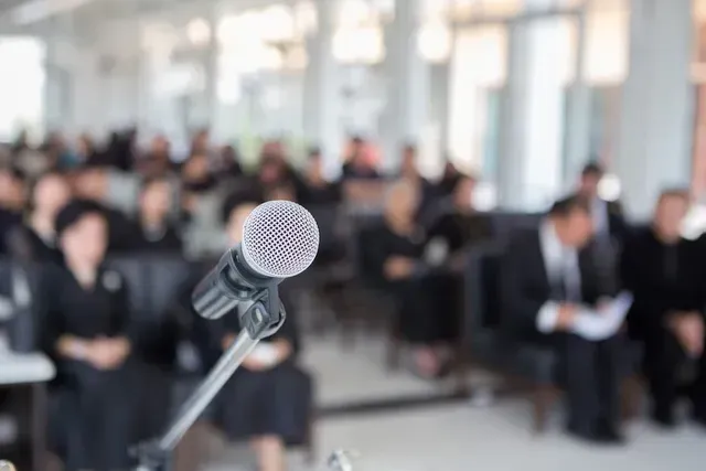 Microphone on stand in foreground with blurred audience seated in a bright conference hall.