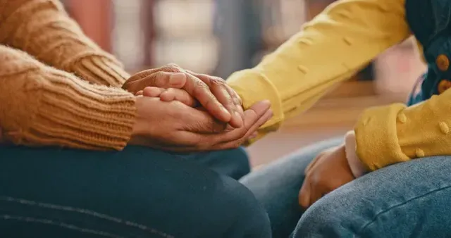 Two people sitting close together, holding hands in a supportive gesture.